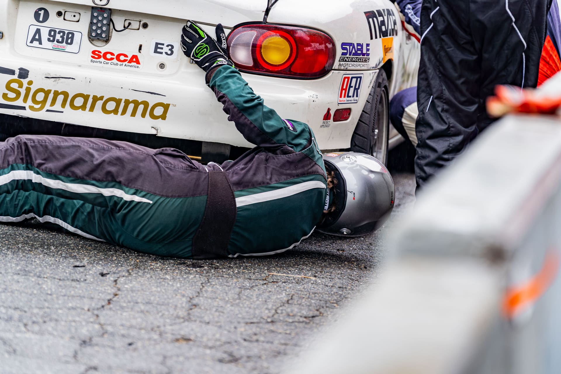 A crew member flat on his back under the rear of the car on wet asphalt.